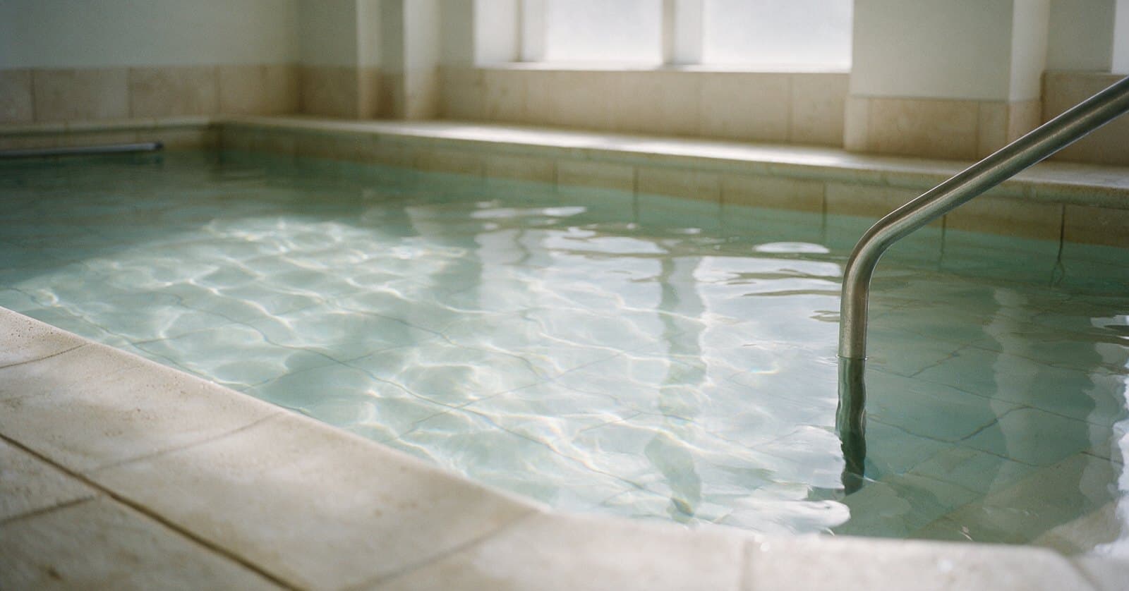 Still life in soft daylight: the pale stone edge of a still indoor therapy pool, refracted light moving across the tiled floor beneath the surface, and a stainless handrail entering the water.